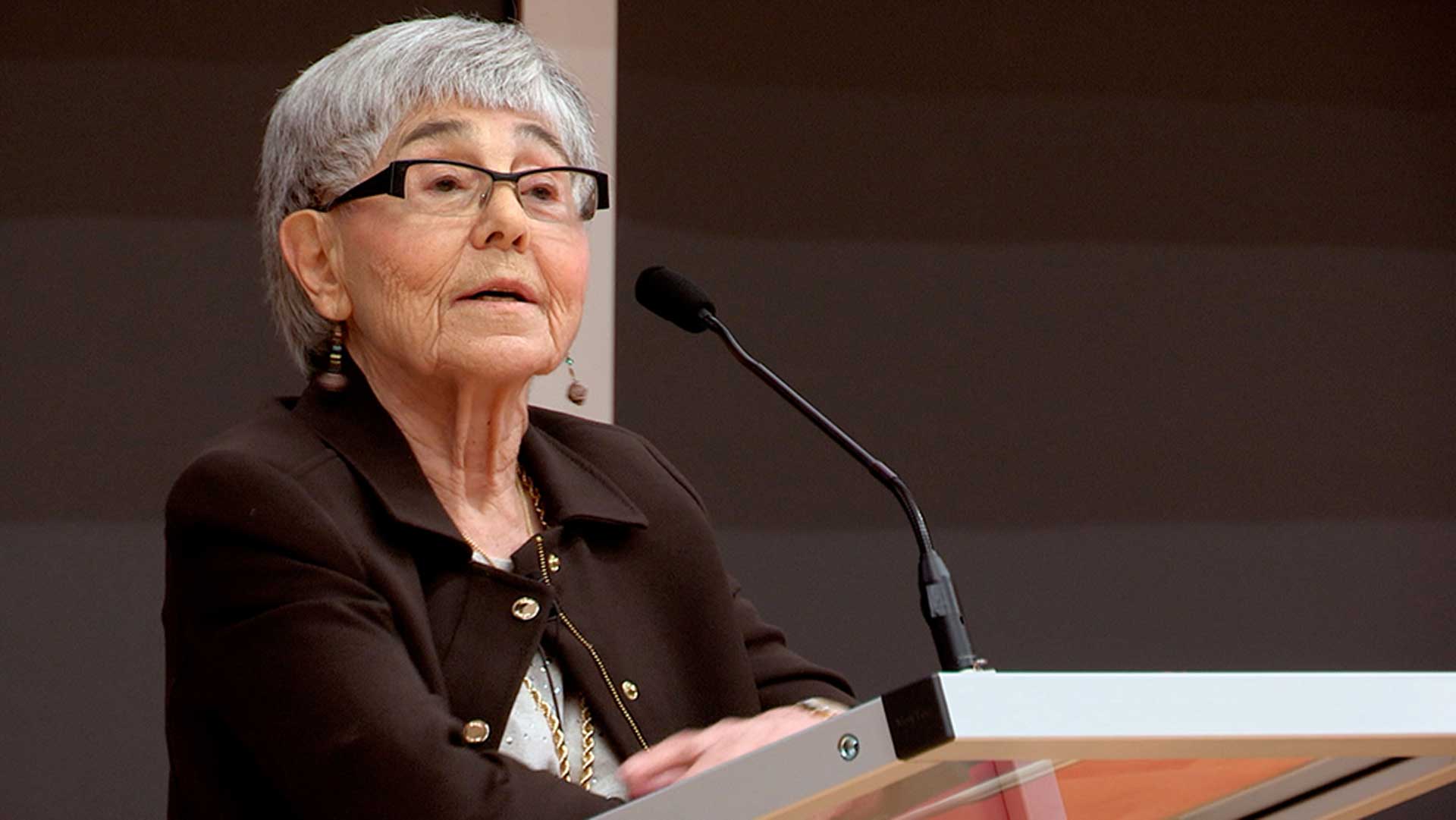May Cohen standing at a podium speaking into a microphone