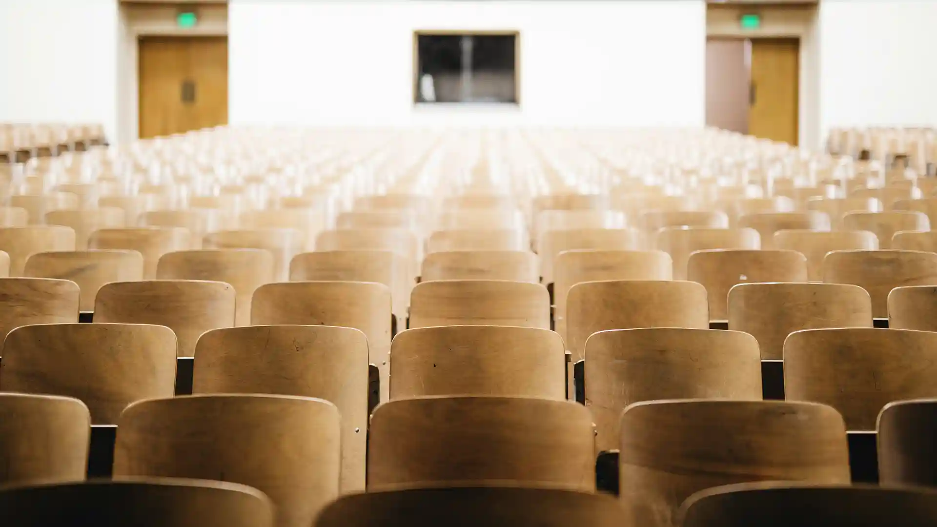 An auditorium of empty wooden seats looking back from the front of the room to the back with two exit doors on either side and a window in the middle.