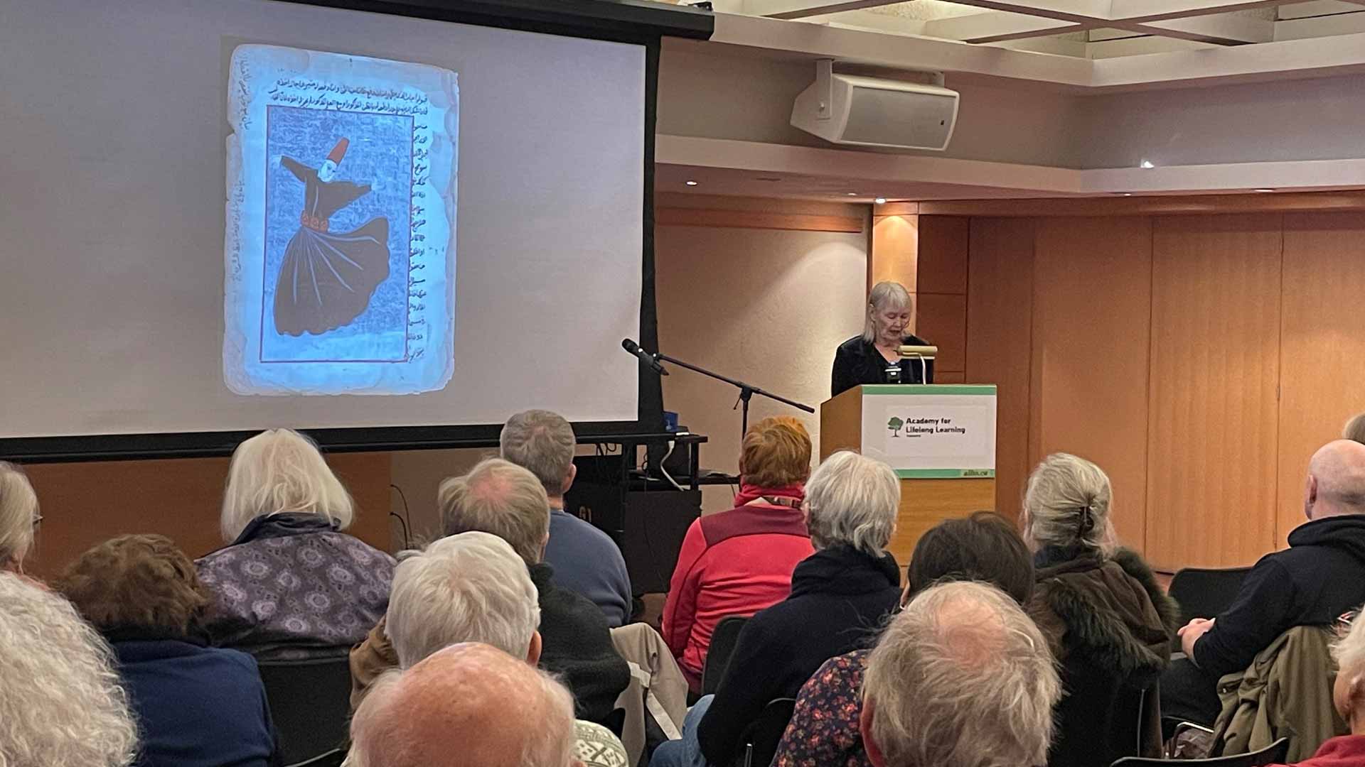 A woman at a podium in a classroom. There is a screen to her right with an image of a piece of art projected on it. An older audience is sitting facing the screen.